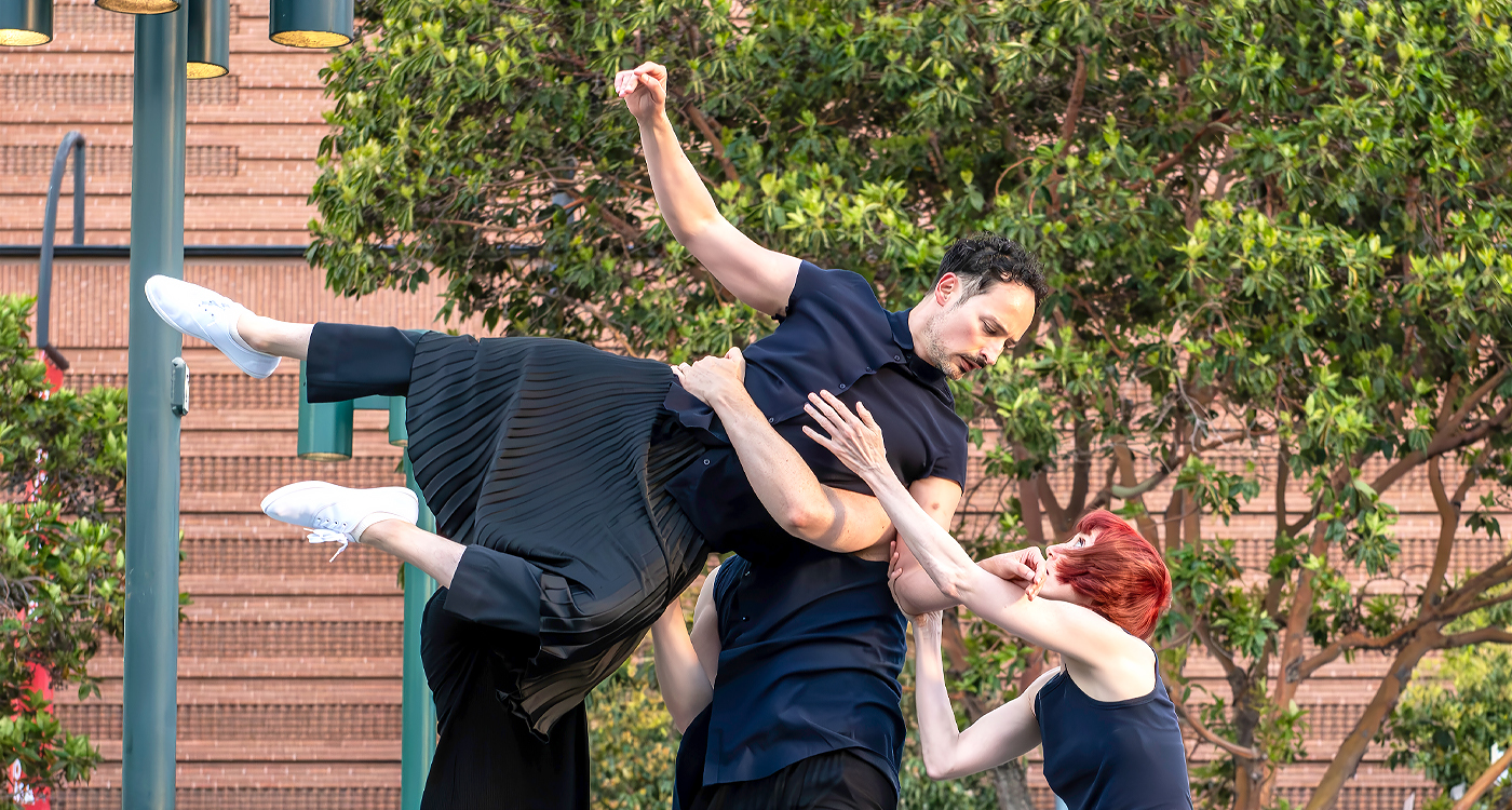 A dancer dressed in black clothes and white sneakers is lifted so their body is horizontal by 3 other dancers dressed in black. The lifted dancer raises one arm and makes eye contact with one of the people lifting them who outstretches their arm to the lifted dancer.