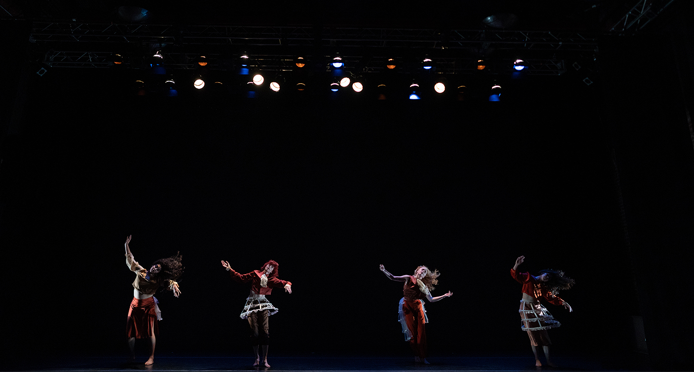 4 female dancers are illuminated on a dimly lit stage, all dressed in various shades of orange and white, and holding the position as though they are partner dancing with an imaginary person.