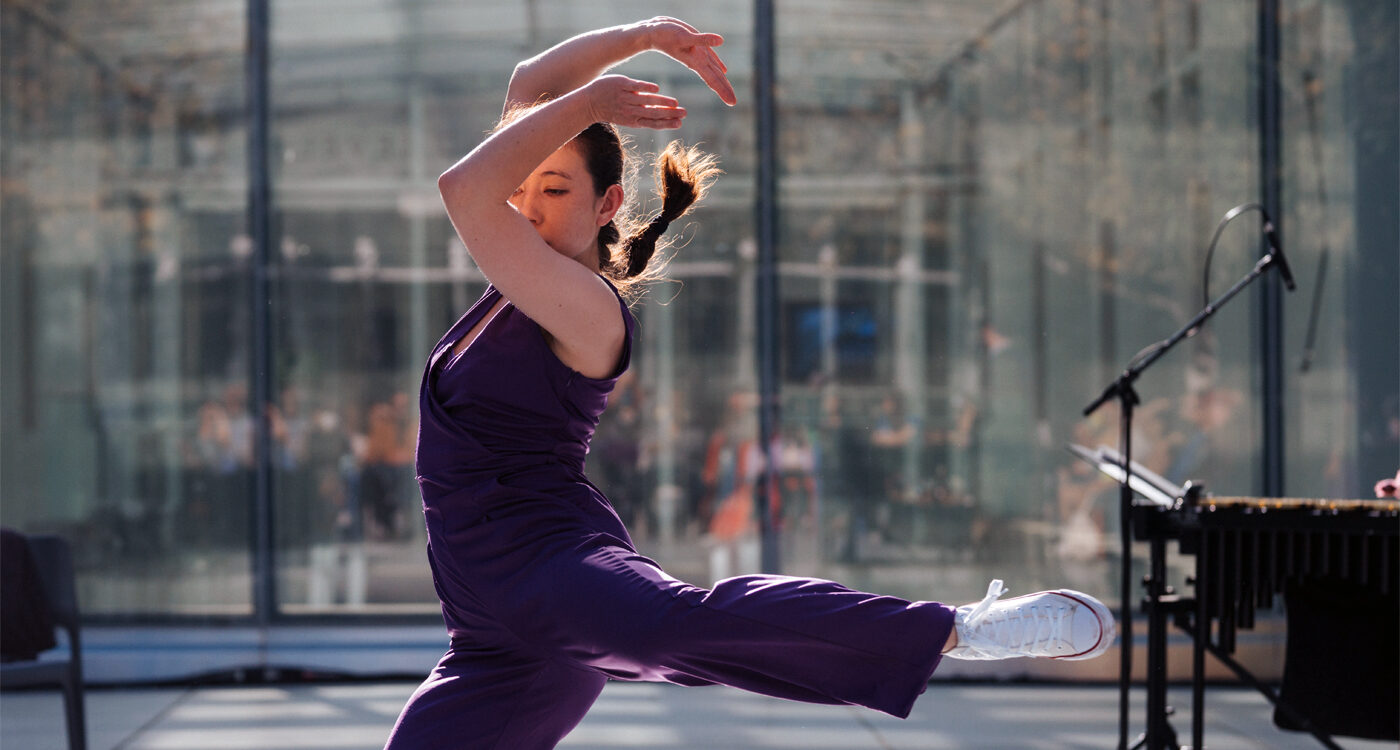 Dancer in a purple jumpsuit kicks leg out to the side with arms above their head