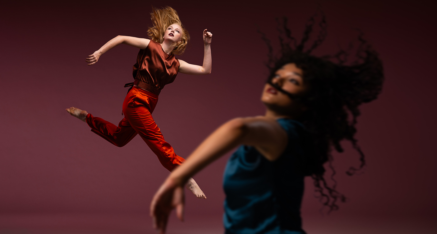 A dancer blurred in the foreground tosses their hair and lifts their arms wearing a blue satin top and another dancer in focus in the background leaps while looking up in an orange and rust satin outfit.