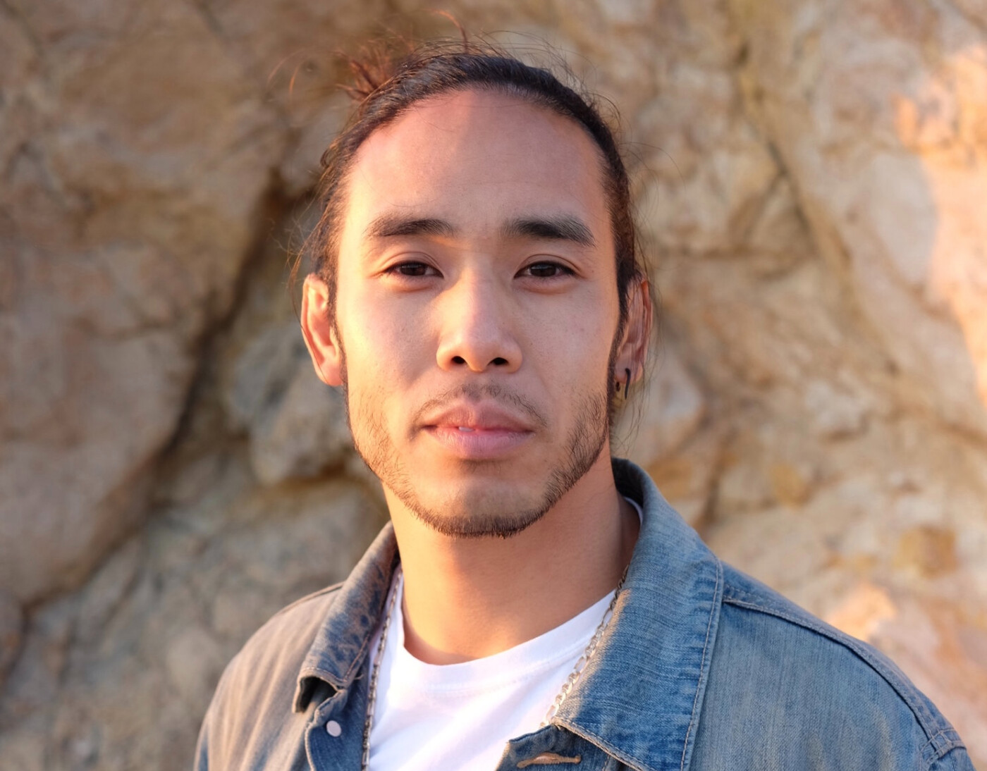 Headshot of Johnny Huy Nguyen, a Vietnamese American man in jean jacket with hair pulled up stares at the camera in front of a stone background.