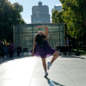 Dancer in a purple dress arches back to look at the camera on one leg in an outdoor area with glass buildings and trees.