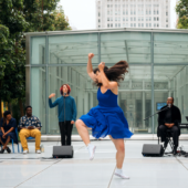 Dancer in a blue dress raises her fists and stomps in front of a live band and spoken word poet outdoors.
