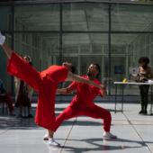 2 dancers in red partner in front of a DJ and a glass building, one dancer leaning back with her leg extended and the other holding her hand.