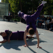 2 dancers in purple outfits perform for an outdoor audience. One dancer is in a handstand while the other is rolling on the ground.
