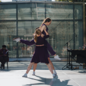 2 dancers lift a third dancer who looks like they’re running in the air. Everyone is in purple in front of a live band and a glass building.