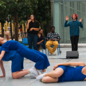 3 dancers in blue outfits perform outdoors in front of a spoken word poet. One dancer is reclining on the ground, one dancer is on their hands and knees while the other is laying across their back.
