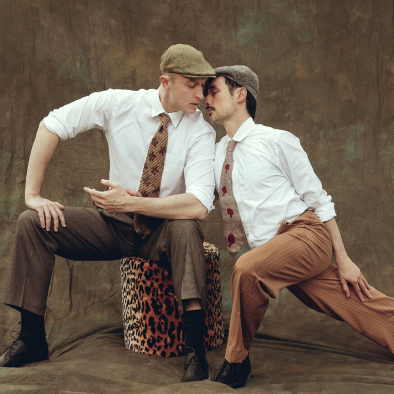 Two male dancers lean into one another, one sitting on a stool and one lunging.
