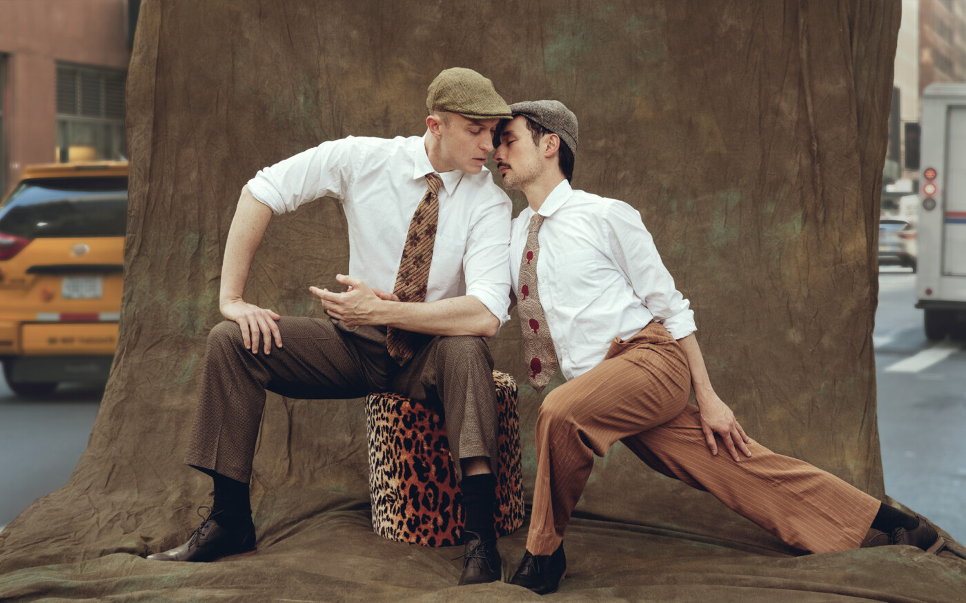 Two male dancers lean into one another, one sitting on a stool and one lunging.