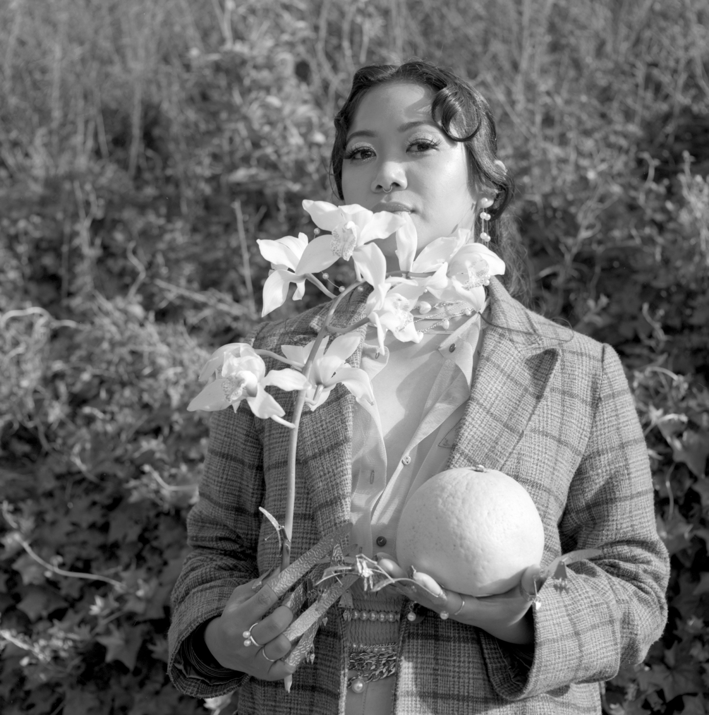 b&w image of Kim, standing against tall grass, holding and orchid plant in one hand hat partially covers her face, and a pomelo in the other. She's wearing a checked coat and has stylized makeup and slicked hair.