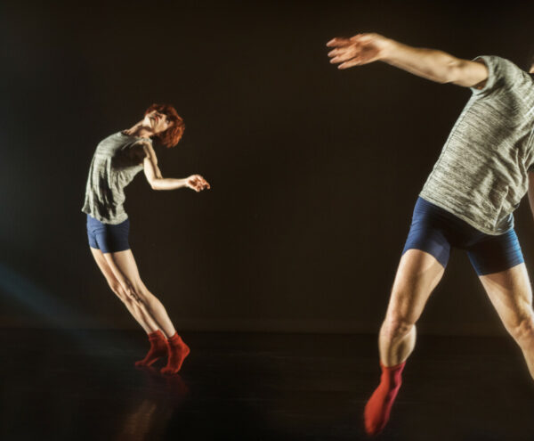 Two dancers caught in suspended movement, both wearing short blue shorts, red socks, and black and white heathered tees. In the foreground, Ryan tilts to the side almost out of the frame. In the back, Wendy is arching back off her weight, looking towards Ryan.