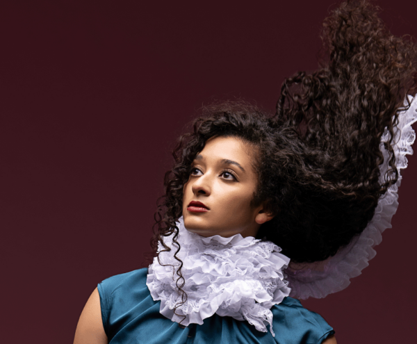 Headshot of a dancer with her gaze lifted to the light and her long, dark curly hair flung high behind her. She's wearing a teal satin top and her neck is wrapped in a long strand of white ruffles that trails, floating with her hair. The background is a rich plum color.