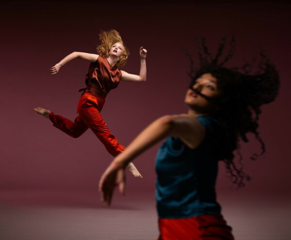 A dancer blurred in the foreground tosses their hair and lifts their arms wearing a blue satin top and another dancer in focus in the background leaps while looking up in an orange and rust satin outfit.