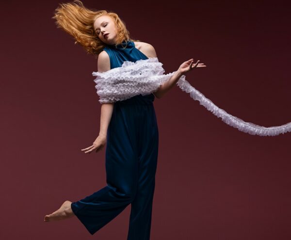 Dancer with flying long strawberry blonde hair looks away from hand while flicking leg behind them. They are wrapped in white ruffle trim in front of a plum background.