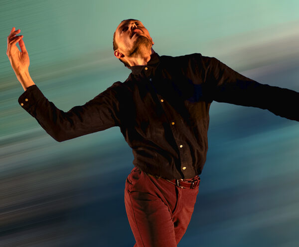 Dancer in a black button up shirt and red pants leans backwards with their arms extended in front of a blurred green background.