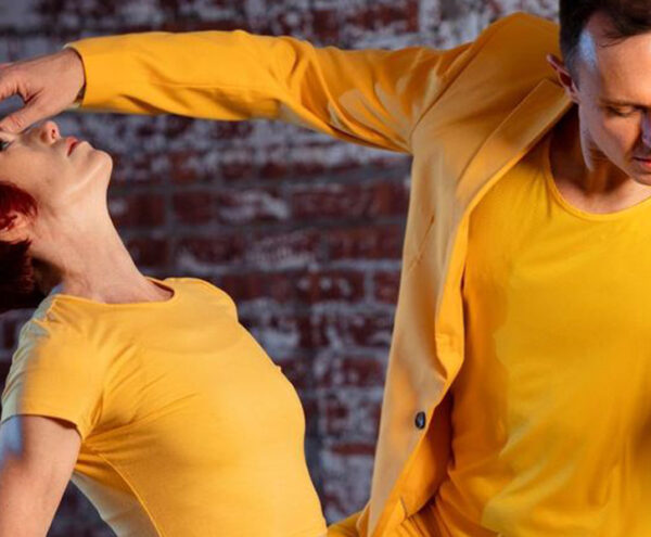 A dancer in a yellow shirt and yellow suit jacket looks down with his arms raised, one arm hovering over the face of another dancer in yellow bending backwards.