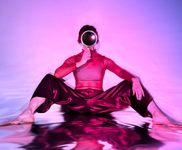 A dancer sits tall on a silvery metallic surface against a saturated pink and purple background. Her bent legs are wide and she's holding a round mirror in front of her whole face. She's wearing a sheer burgundy top and shiny burgundy pants. The floor has a watery feel, distorting her reflection.