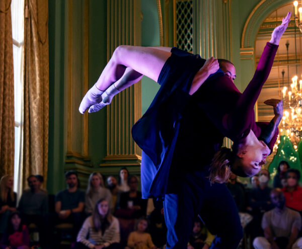 One dancer lifts another over their shoulder. The lifted dancer is in a back bend with one arm extended. The dancers are in a golden and green ornate room.